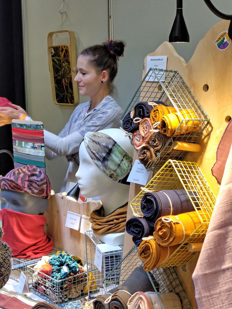 Frau hinter einem Verkaufsstand und überreicht ein Produkt an eine Kund:in. Diese Person ist jedoch nicht auf dem Bild. Im Vordergrund ist ein Regal auf dem Verkaufsstand, in dem Reisehandtücher ausgestellt sind.
