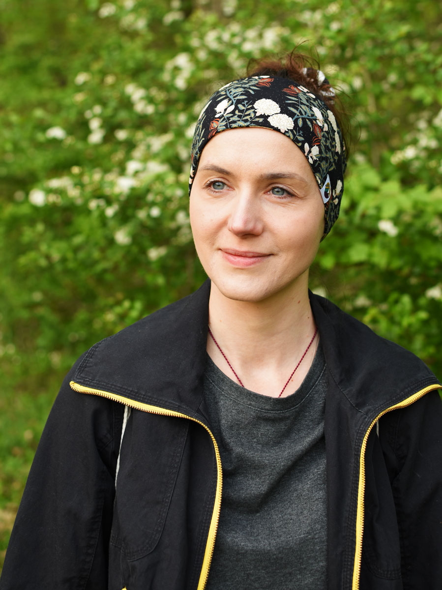 Porträt einer Frau mit gemustertem Stirnband und schwarzer Jacke vor grünem, unscharfem Waldhintergrund