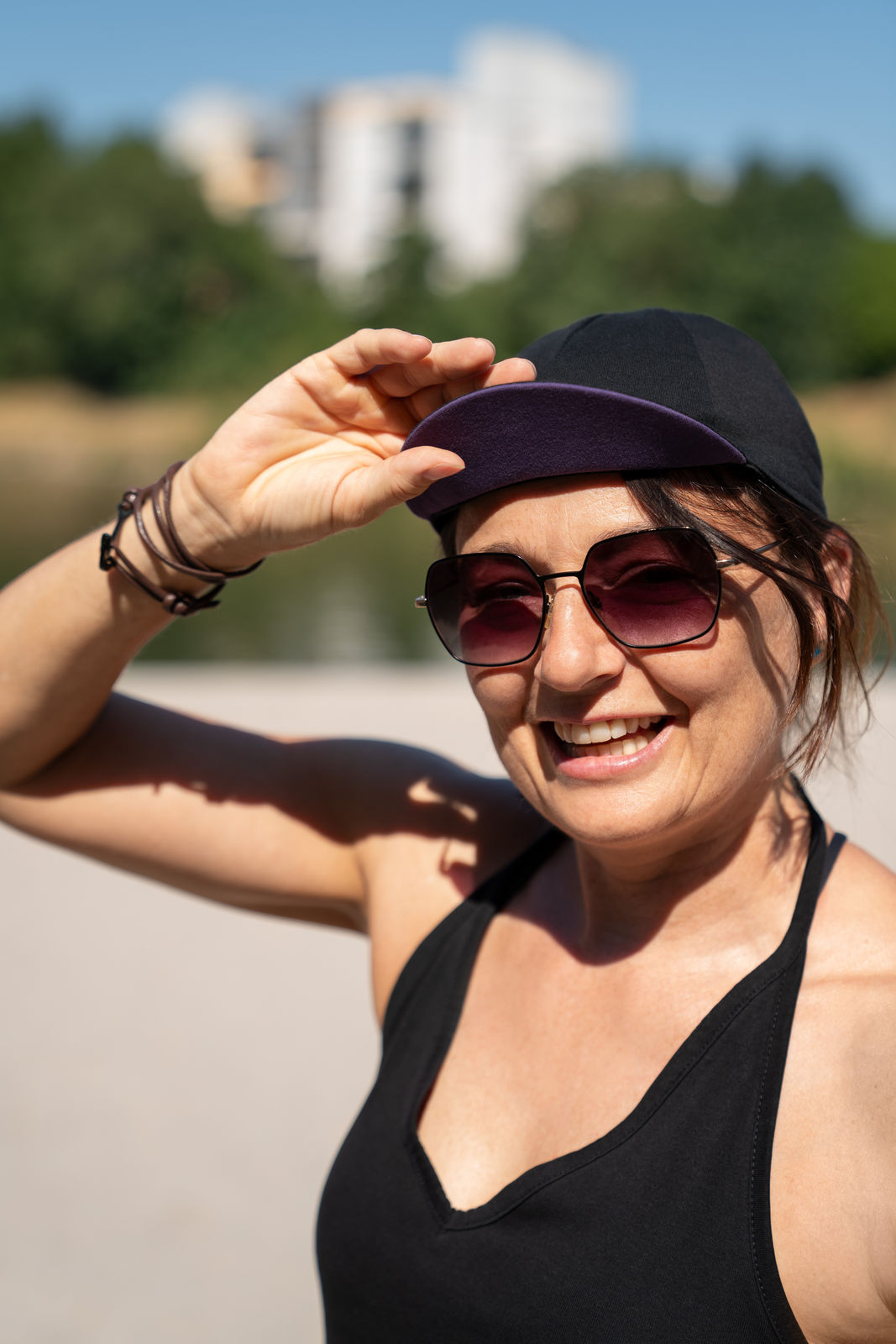 Lächelnde Frau am Strand mit Sonnenbrille und Fahrrad Cap hebt die Hand an das Mützenschild, Hintergrund unscharf mit Landschaft und Gebäuden.
